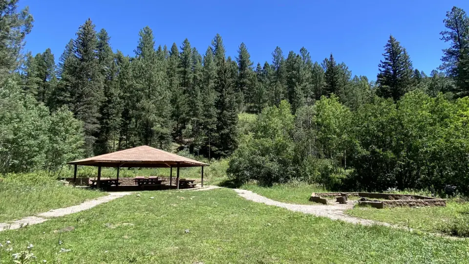 A ramada next to campfire is surrounded by conifers and aspen in a sunny meadow.