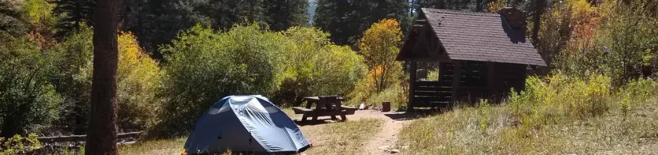 A sheltered site with a tent, picnic table, and fire place with fall foliage and pines in the background.