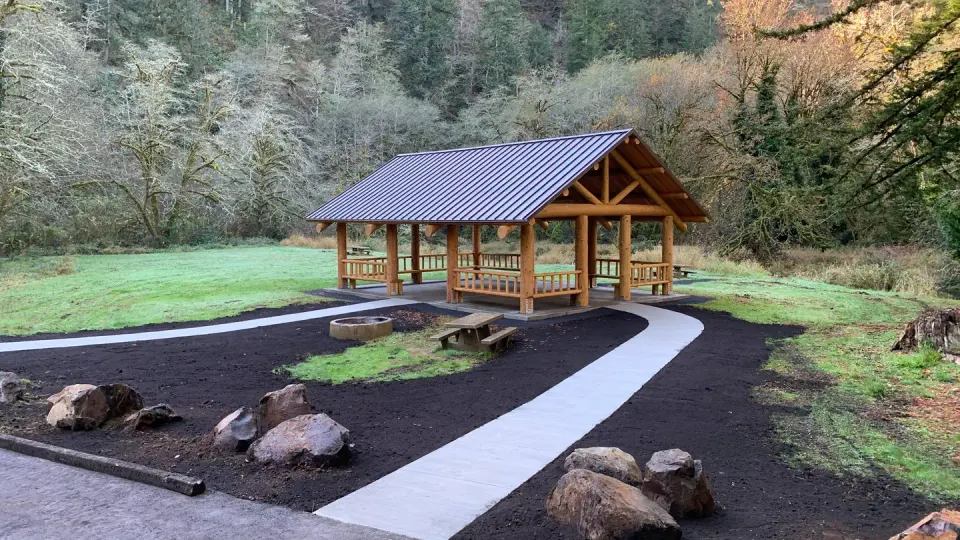 Picnic shelter. Large flat field. Tents and picnic tables in front of a bridge, a large conifer tree, and mixed conifer forest in the back, fire ring and picnic table behind single pole fence in foreground.